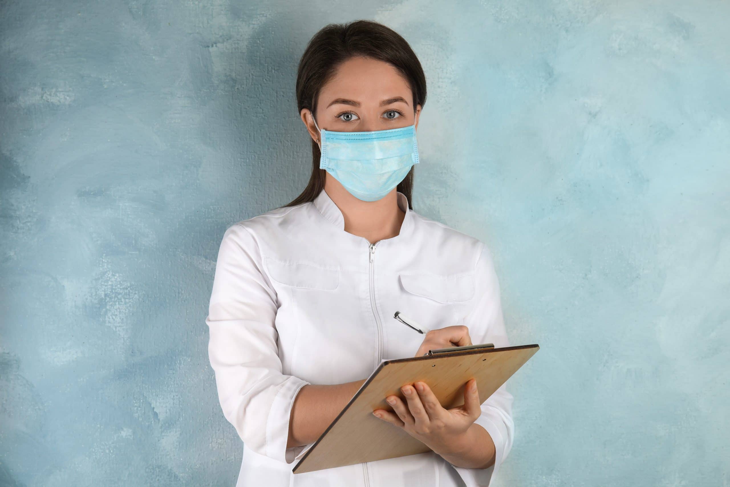 A doctor working in a long term care facility wears a disposable mask and takes notes on a clipboard.Behind her is a light blue wall.