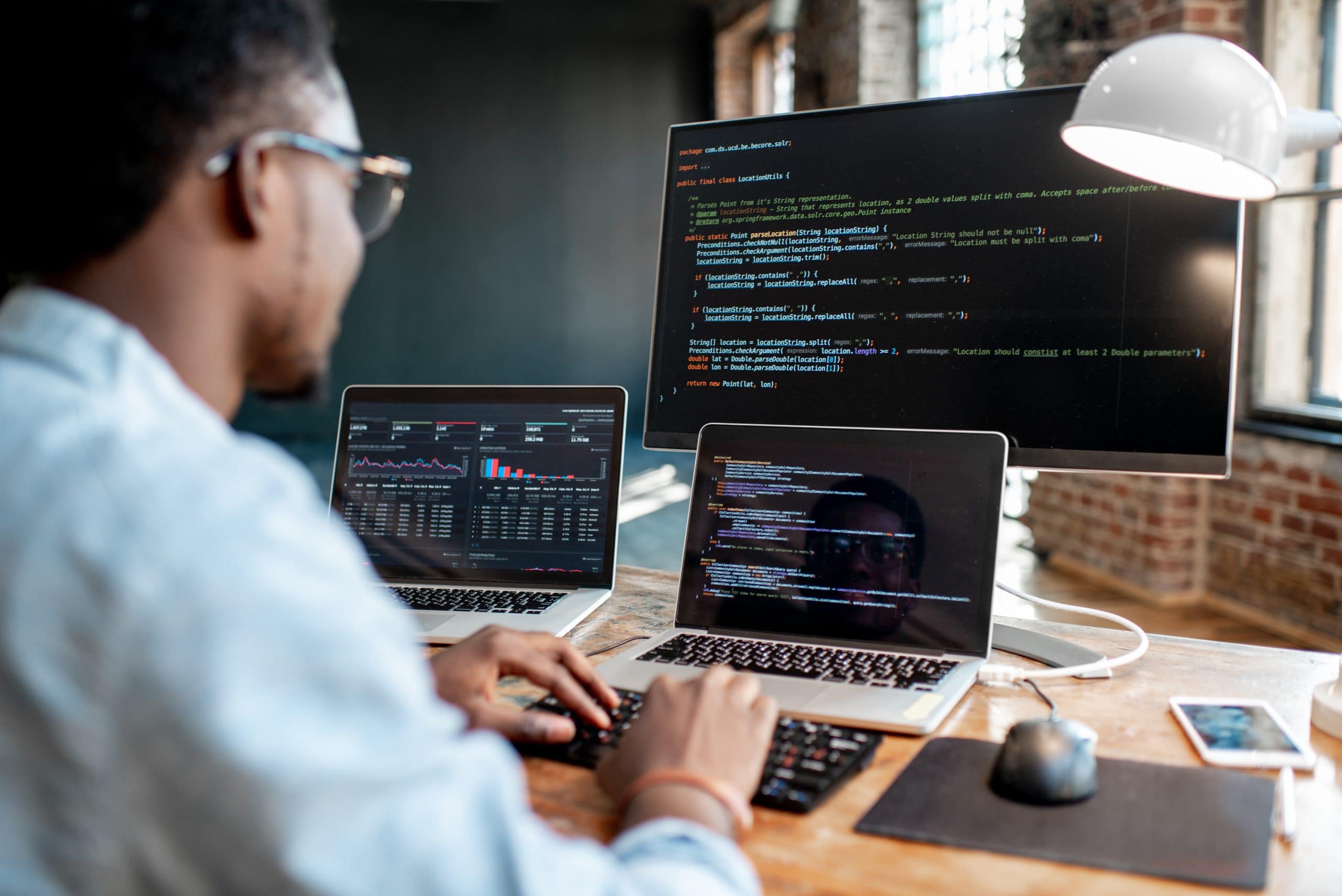 Young Black male programmer writing program code sitting at the workplace with three monitors in the office. Image focused on the screen