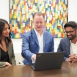 Three team members at a management consulting firm work together on a laptop. Behind them is a colourful abstract painting.