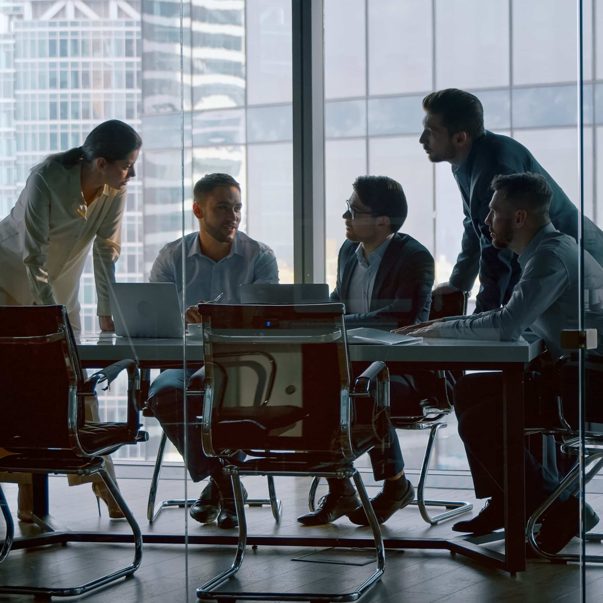 Five team members collaborate around a table in a meeting room with a wall of windows