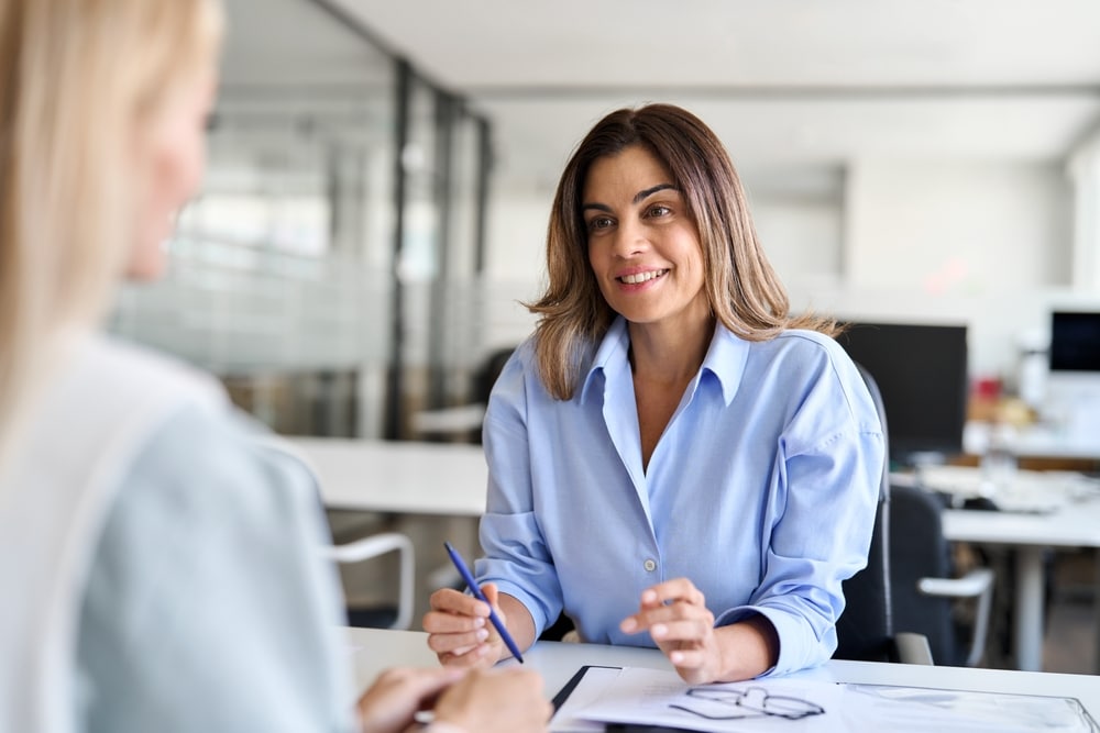 2 women talking over a desk with papers on it in an office setting.