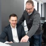 A man standing over another man sitting at a desk pointing at a computer screen.