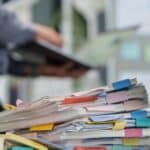 tressed employee hand holding a stack of disorganized documents and files with colorful binder clips on a chaotic office desk, experiencing business burnout and pressure from work overload