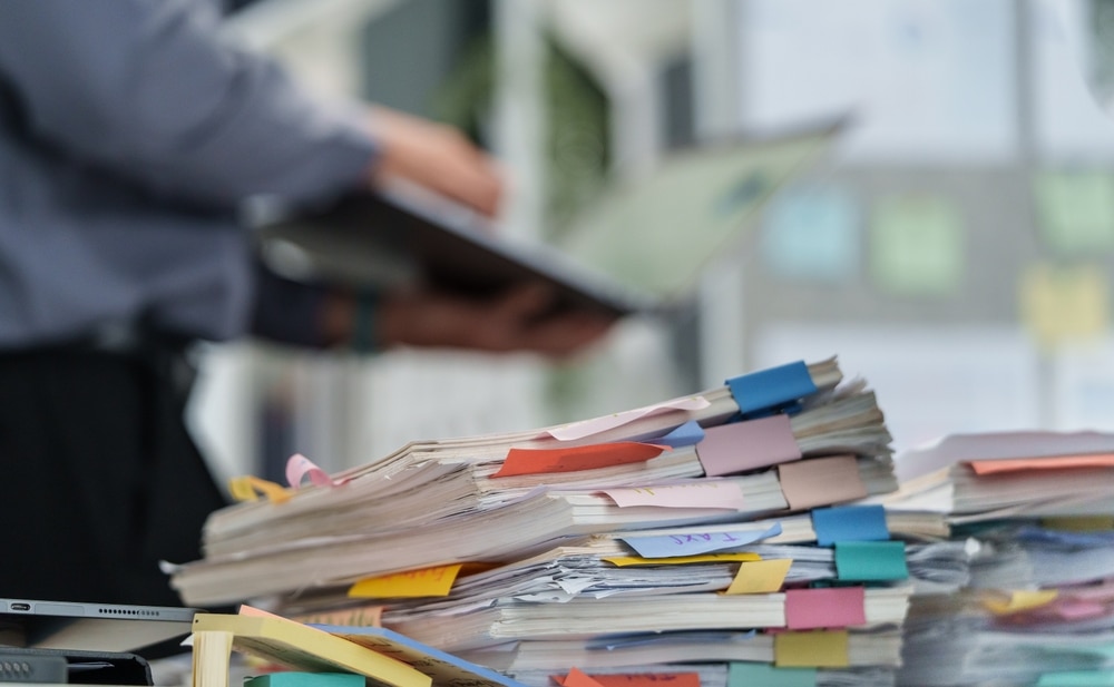 tressed employee hand holding a stack of disorganized documents and files with colorful binder clips on a chaotic office desk, experiencing business burnout and pressure from work overload